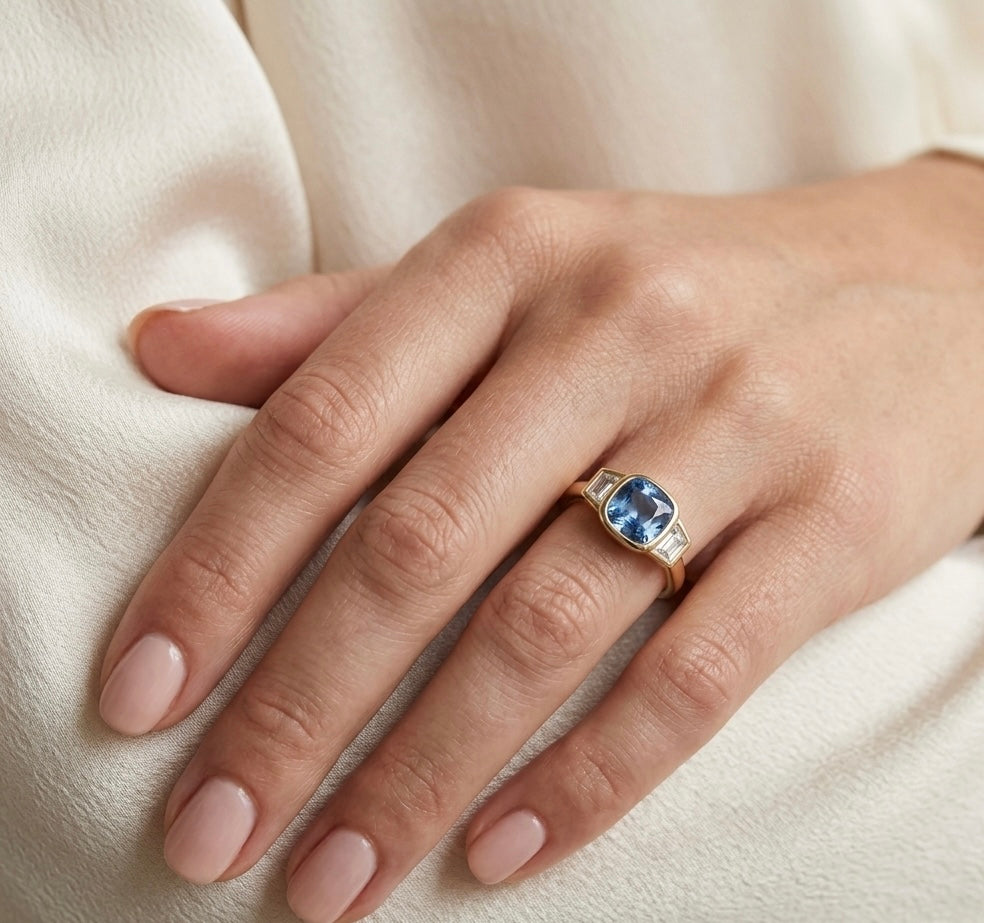 Hand wearing a gold ring with a blue gemstone on a beige fabric background. Gold cast in Penzance, Cornwall. 